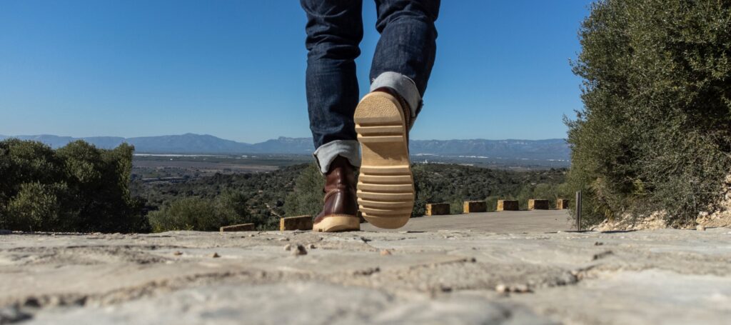 Person geht auf einem sonnigen Weg in einer natürlichen Landschaft spazieren. Die Aufnahme zeigt die Beine und Schuhe aus niedriger Perspektive, im Hintergrund sind Bäume, eine weite Ebene und Berge zu sehen.